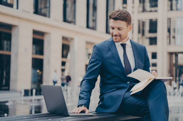 Professioneller junger Mann im blauen Anzug, der am Laptop recherchiert oder ein Business-Webinar besucht, wichtige Informationen in Notizbüchern notiert und mit gekreuzten Beinen auf einer Bank sitzt - Foto, Bild