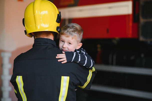 Dirty firefighter in uniform holding little saved boy standing on black background - Foto, Imagem