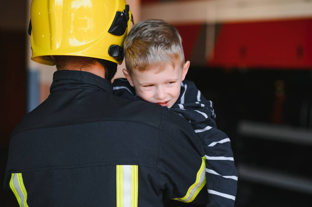 Dirty firefighter in uniform holding little saved boy standing on black background - Fotografie, Obrázek