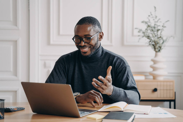 Afroamerykanin młody biznesmen w okularach mający konferencję online z pracodawcami za pośrednictwem połączenia wideo szeroko uśmiechnięty patrząc na ekranie laptopa gestykulując podczas komunikacji i odpowiadając na pytania - Zdjęcie, obraz