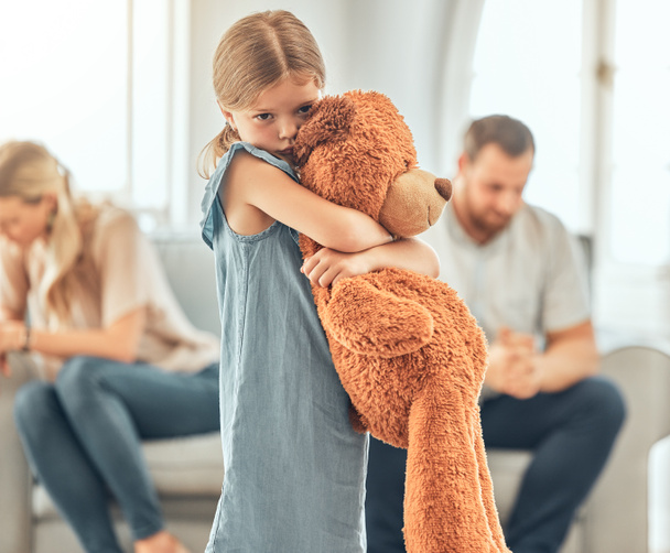 An upset little girl squeezing her teddy bear while looking sad and depressed while her parents argue in the background. Thinking about her parents breaking up or getting divorced is causing stress. - Foto, imagen