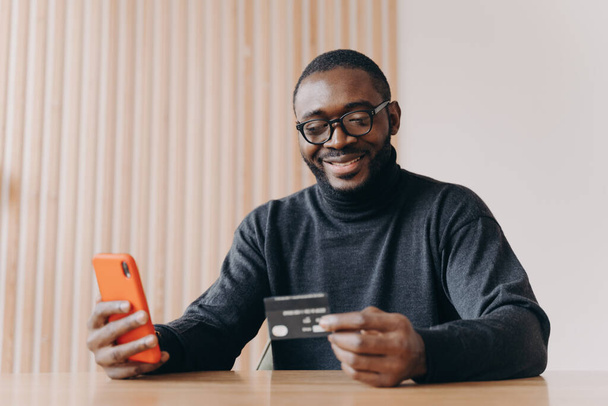 Cheerful Aframerican man entrepreneur holding credit card and using touchscreen smartphone for online shopping while making orders online sitting at home office. Ecommerce and online payment concept - Foto, imagen