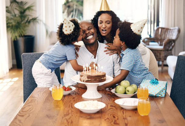 Africká americká rodina nosí klobouky a doma slaví narozeniny s dortem. Malí sourozenci objímají a líbají svého otce na jeho narozeniny. Šťastný muž se usmívá po narozeninovém překvapení. - Fotografie, Obrázek