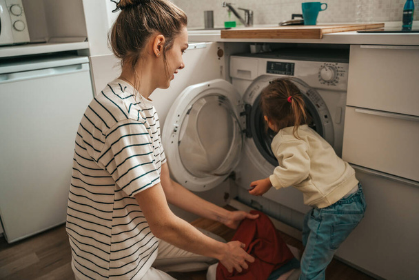 Child placing the laundry items into the open washing machine assisted by her female parent - 写真・画像