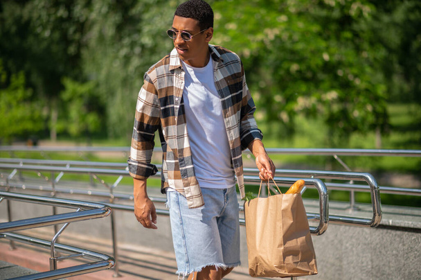 After shopping. Young dark-skinned man in plaid shirt walking with a paper bag in hands - Foto, Imagen