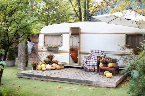 Campsite in garden. Halloween design home. Interior cozy yard campsite with fall flowers potted and pumpkins. Wooden RV house porch with garden furniture. Wooden armchair near outside caravan trailer  - Foto, immagini