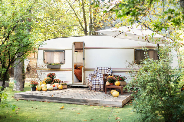 Halloween design home. Interior cozy yard campsite with fall flowers potted and pumpkins. Wooden RV house porch with garden furniture. Campsite in garden. Wooden armchair near outside caravan trailer  - Фото, изображение