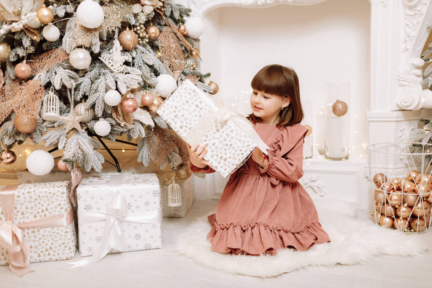 Rozkošná holčička s dárky, dárkové krabice u vánočního stromečku. Usmívající se dítě v růžových šatech u zdobeného krbu. Happy family at home Christmas New Year December time for celebration concept. - Fotografie, Obrázek