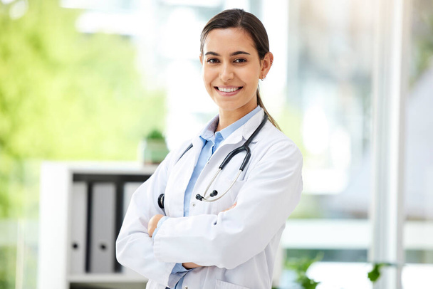 Confident young mixed race female doctor standing with her arms crossed inside a medical office. One hispanic woman in a white coat with stethoscope. Trusted practitioner caring for the health of pat. - Foto, imagen