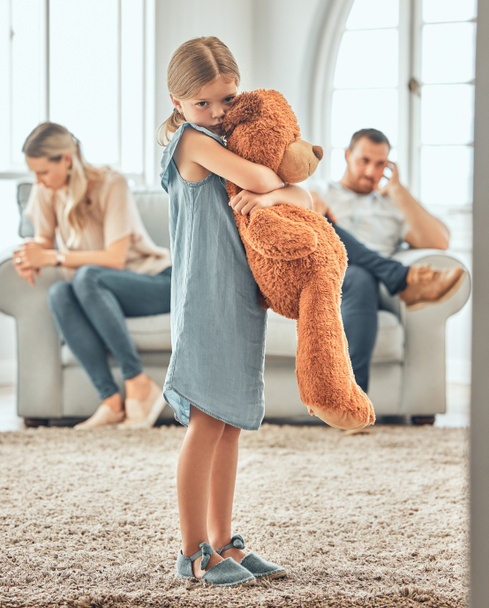An upset little girl squeezing her teddy bear while looking sad and depressed while her parents argue in the background. Thinking about her parents breaking up or getting divorced is causing stress. - Фото, изображение