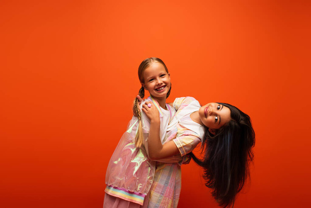 veselá dívka objímající brunetka přítel při úsměvu na kameru izolované na oranžové - Fotografie, Obrázek