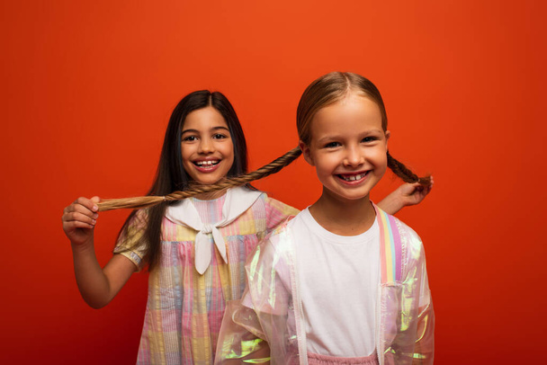 smiling girl holding pigtails of happy friend looking at camera isolated on orange - Φωτογραφία, εικόνα