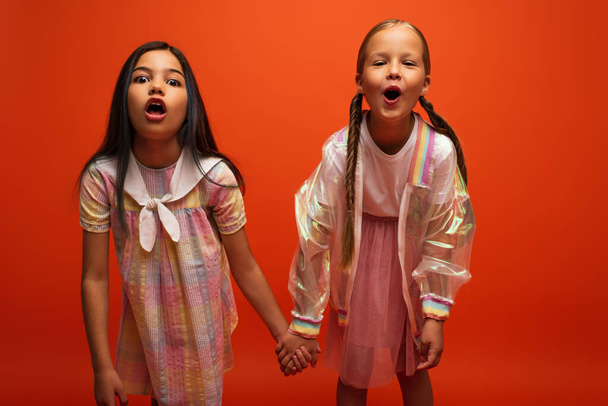 grappige meisjes met open monden hand in hand en grimmig op camera geïsoleerd op oranje - Foto, afbeelding