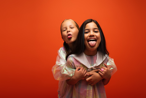 excited friends embracing and sticking out tongues while having fun isolated on orange - Photo, Image