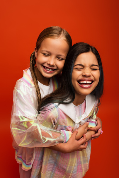 overjoyed girls hugging and laughing with closed eyes isolated on orange - Photo, Image