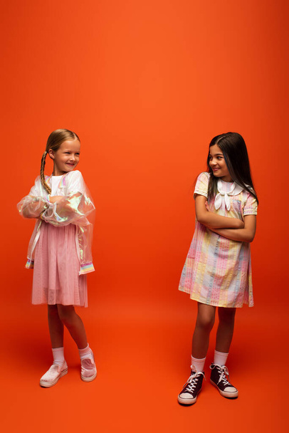 full length of cheerful preteen girls standing with crossed arms and smiling at each other on orange background - Foto, afbeelding