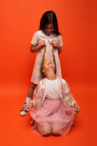 happy girl sitting and looking at friend holding her pigtails on orange background - Foto, imagen