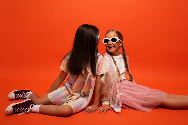 stílusos tizenéves gyerekek napszemüvegben mosolyognak egymásra, miközben ül a narancssárga háttér - Fotó, kép