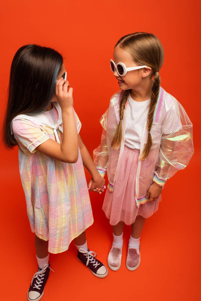 high angle view of girls in trendy clothes and sunglasses holding hands and smiling at each other on orange background - Foto, Imagen