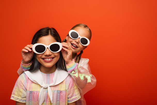 cheerful kid putting sunglasses on smiling brunette girl in dress isolated on orange - Photo, Image