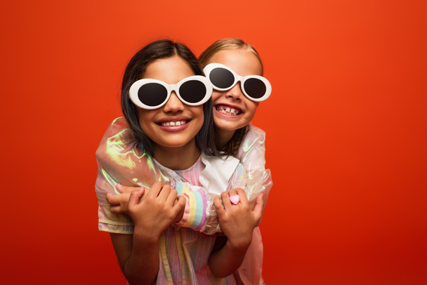 filles heureuses dans des lunettes de soleil élégantes étreignant et souriant isolé sur orange - Photo, image