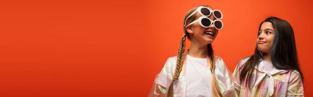 excited girl in two sunglasses having fun near brunette friend isolated on orange, banner - Valokuva, kuva