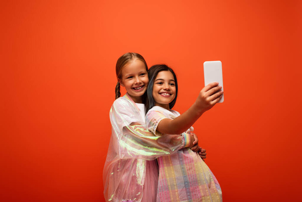girl in rain jacket hugging friend in dress taking selfie on cellphone isolated on orange - Photo, Image