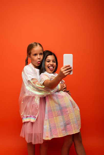 cheerful preteen girls sticking out tongues while taking selfie on mobile phone isolated on orange - Foto, immagini