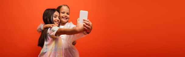 улыбающиеся девочки-подростки в стильной одежде, обнимающиеся и делающие селфи на смартфоне, изолированные на оранжевом, баннер - Фото, изображение