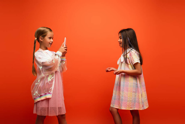 zijaanzicht van brunette meisje gebaren en praten in de buurt vriend opname video op smartphone geïsoleerd op oranje - Foto, afbeelding