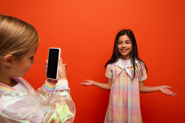 girl with mobile phone taking photo of smiling brunette friend posing with open arms isolated on orange - Photo, Image