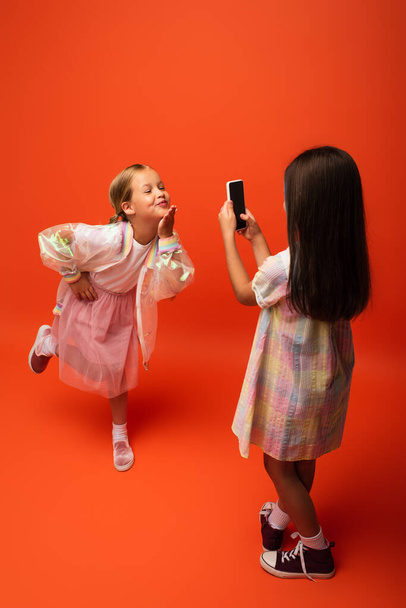 veselá dívka pózuje s rukou na boku a foukání vzduchu polibek v blízkosti přítele fotografování na mobilu na oranžovém pozadí - Fotografie, Obrázek