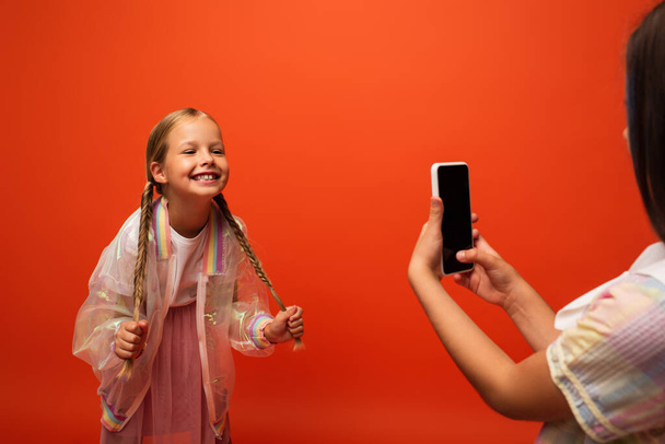 友達の近くで彼女のピグテールを持っている面白い女の子はオレンジで隔離されたスマートフォンで写真を撮る - 写真・画像