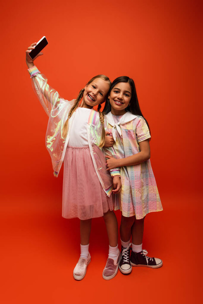 teljes hossza boldog lány esőkabát csinál szelfi barátjával ruha narancs háttér - Fotó, kép