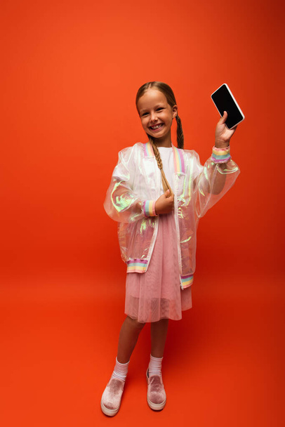 πλήρες μήκος του χαρούμενου κοριτσιού αγγίζοντας κοτσίδα και δείχνει smartphone με κενή οθόνη σε πορτοκαλί φόντο - Φωτογραφία, εικόνα