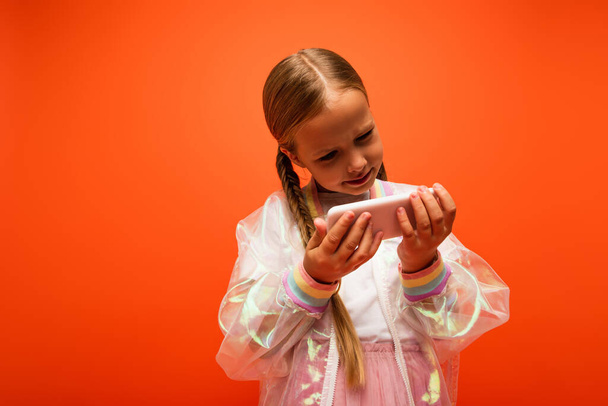 girl in transparent rain jacket looking at mobile phone isolated on orange - Photo, Image