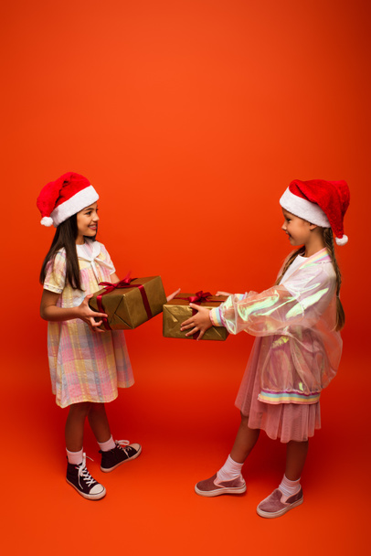 full length of smiling preteen girls in santa hats holding christmas presents on orange background - Фото, изображение