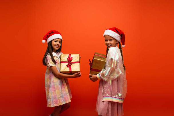 cheerful girls in santa hats holding gift boxes and smiling at camera isolated on orange - Fotografie, Obrázek