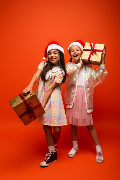 full length of trendy girls in santa hats posing with christmas presents on orange background - Фото, зображення