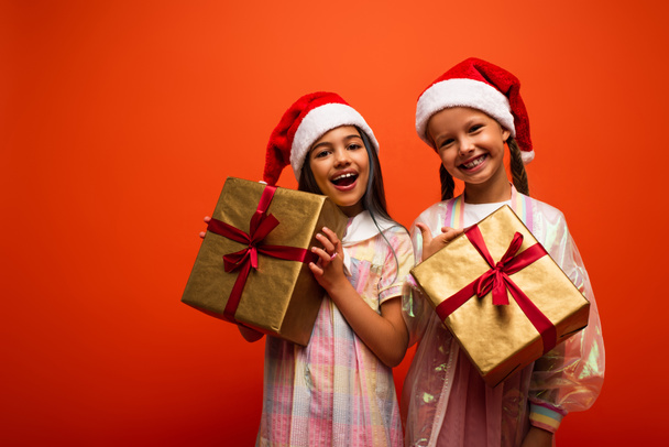amazed and happy girls in santa hats standing with gift boxes and looking at camera isolated on orange - Foto, Bild