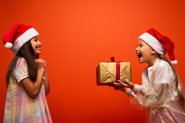 widok z boku zdumiony dziewczyna w Santa kapelusz gospodarstwa prezent świąteczny blisko radosny przyjaciel odizolowany na pomarańczowy - Zdjęcie, obraz