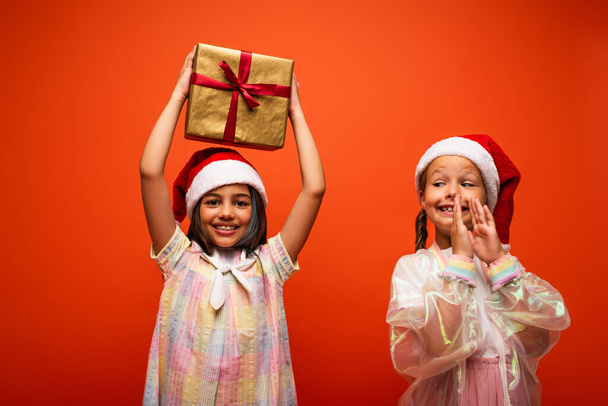 vzrušené dítě v Santa klobouk tleskání rukou v blízkosti dívky s dárkovou krabicí ve zvýšených rukou izolovaných na oranžové - Fotografie, Obrázek