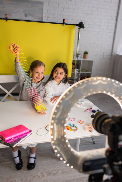 улыбающаяся девушка, показывающая спиральную игрушку, сидя рядом с другом и размытая цифровая камера в круговой лампе - Фото, изображение