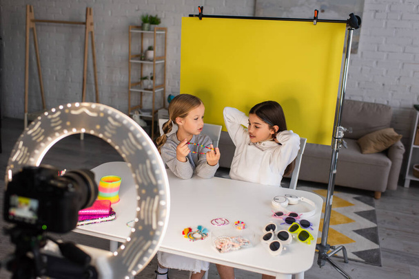 meisje tonen kralen in de buurt van video blogger op gele achtergrond in de voorkant van wazig digitale camera met cirkel lamp - Foto, afbeelding