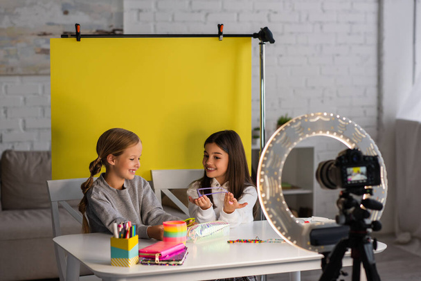 cheerful preteen girls showing sunglasses while recording video blog on blurred digital camera near yellow background - Foto, afbeelding