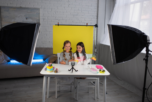 улыбающиеся видеоблоггеры, показывающие спиральную игрушку перед сотовым телефоном на телефоне, рядом с желтым фоном дома - Фото, изображение