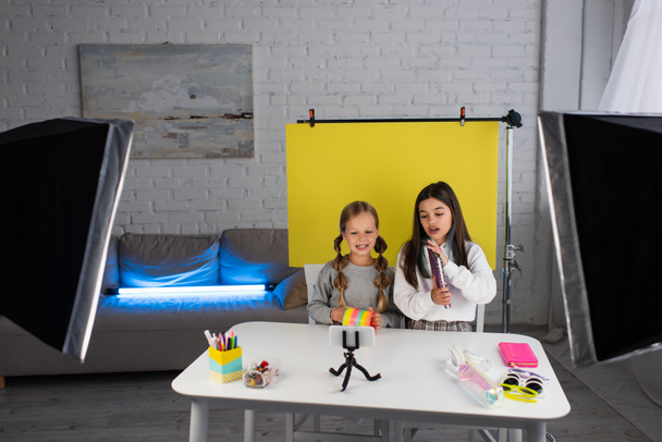 blogger tonen copybook in de buurt van vriend met spiraalvormig speelgoed terwijl zitten op een gele achtergrond in de buurt van smartphone en lampen - Foto, afbeelding
