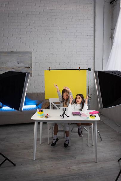 excited girls gesturing while recording video blog on mobile phone near lamps and yellow background at home - Fotografie, Obrázek