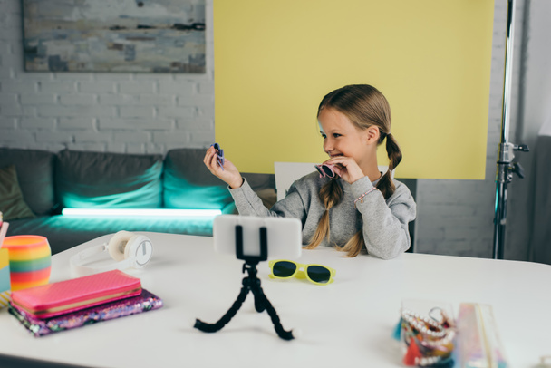 счастливый блоггер показывает стильные солнцезащитные очки возле размытых мобильных телефонов и пенал с наушниками на столе - Фото, изображение