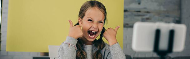 astonished girl with open mouth showing thumbs up near blurred cellphone on yellow background at home, banner - Photo, Image
